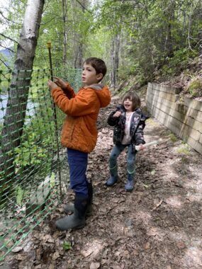 A young boy in a bright, orange jacket attaches green mesh fencing to rebar on a nature trail in the woods. His younger sister stands near him watching with enthusiasm. Below them, through the trees, a river is partially visible. It's a sunny day, so the sunlight casts shadows from the treetops onto the dirt trail.