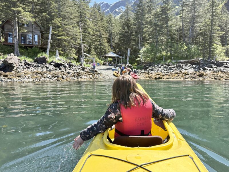 A vibrant shot of a young girl in the front of a yellow kayak with an orange life vest on. She's dipping her left hand into crystal waters as the kayak heads to a shore covered in rocks. Behind that are tall, majestic spruce trees, and behind those, towering mountains with snow still visible on the peaks. A wooden cabin is slightly hidden by the trees. The photo was taken by the person sitting behind the girl in the kayak.