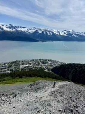 A spectacular vertical photo of a high vantage point looking down on a distant town from the top of a mountain. The scenery is so vast that a person in the foreground even looks tiny. Below is a town next to an azul body of water, and beyond that are snow-covered mountains. The sky is the same color of blue, with white clouds fanning out. The color composition of the photos, with rugged terrain, followed by patches of green grass, then the blue of the water, mountains, and sky, is noteworthy.